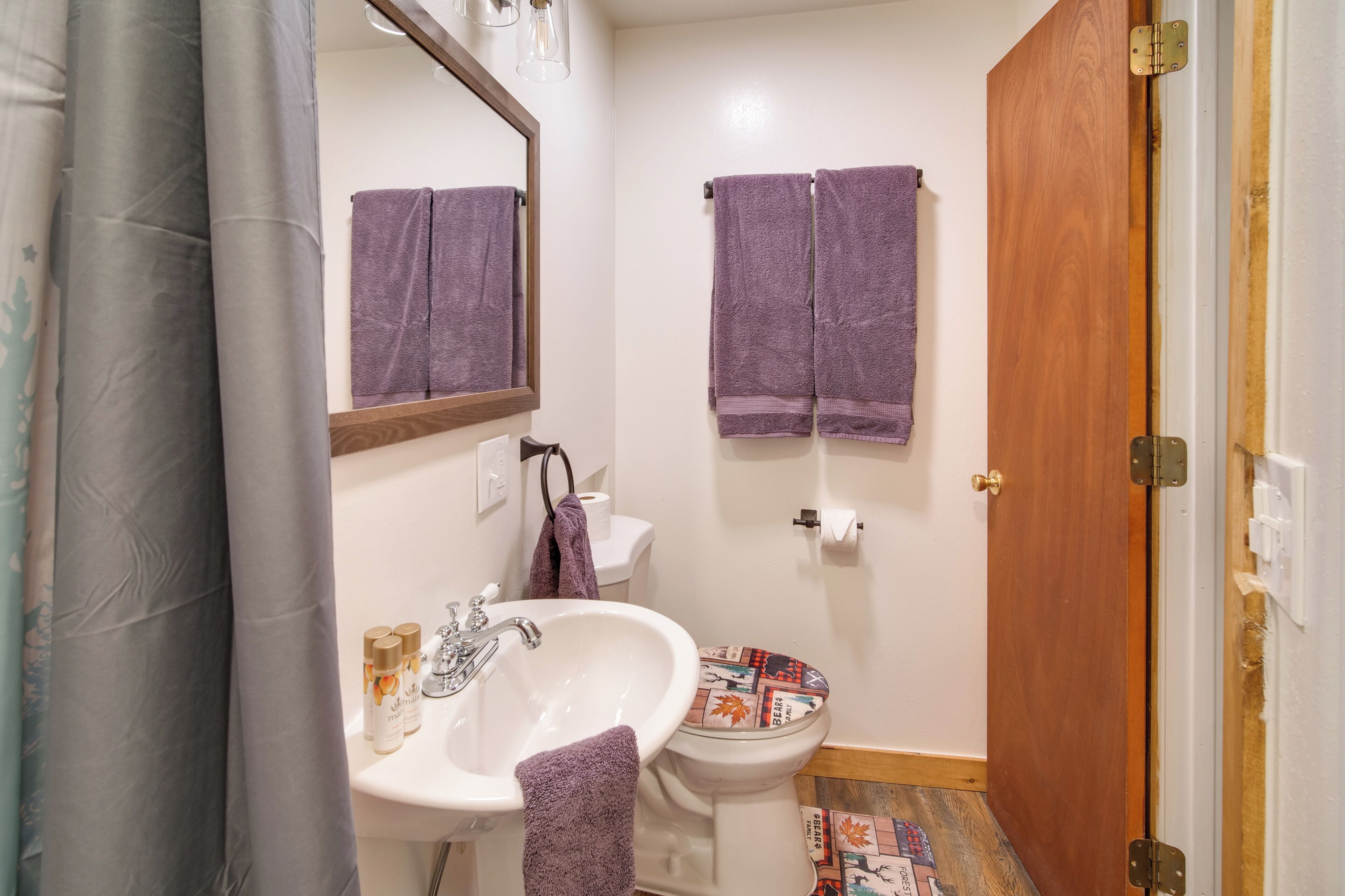 Un baño con un lavabo blanco, un espejo, un inodoro con el asiento estampado y dos toallas moradas colgadas en la pared.
