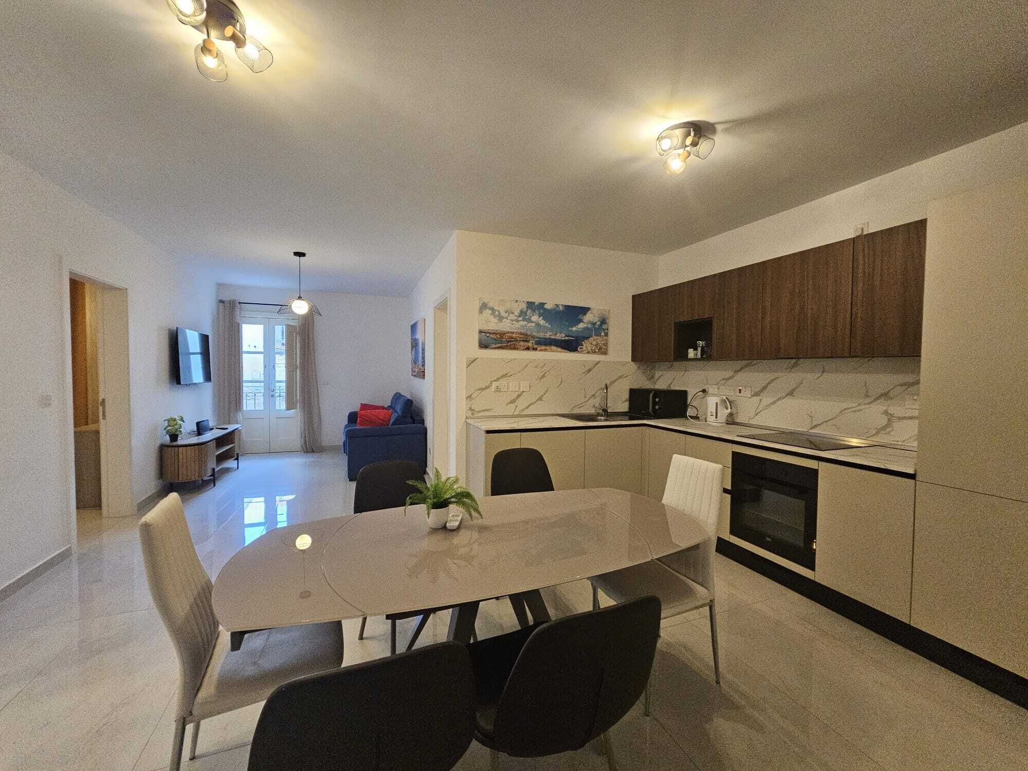 A modern kitchen with a dining area, a TV mounted on the wall, and a painting above the cabinets.