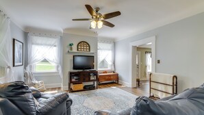 A living room with a ceiling fan, a flat-screen TV, a wooden cabinet, and a large window with sheer curtains. - 'grandma Kellys' Southeast Indiana Farmhouse! (Cedar Grove)