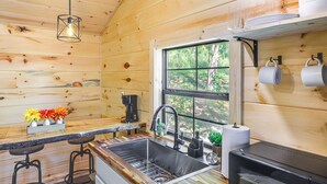Cocina de cabaña de madera con un lavabo, taburetes de bar, una cafetera y una ventana con vistas a árboles.