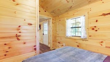 Una habitación de cabaña de madera con una cama, un ventanal con cortinas y una puerta que da a otra habitación.