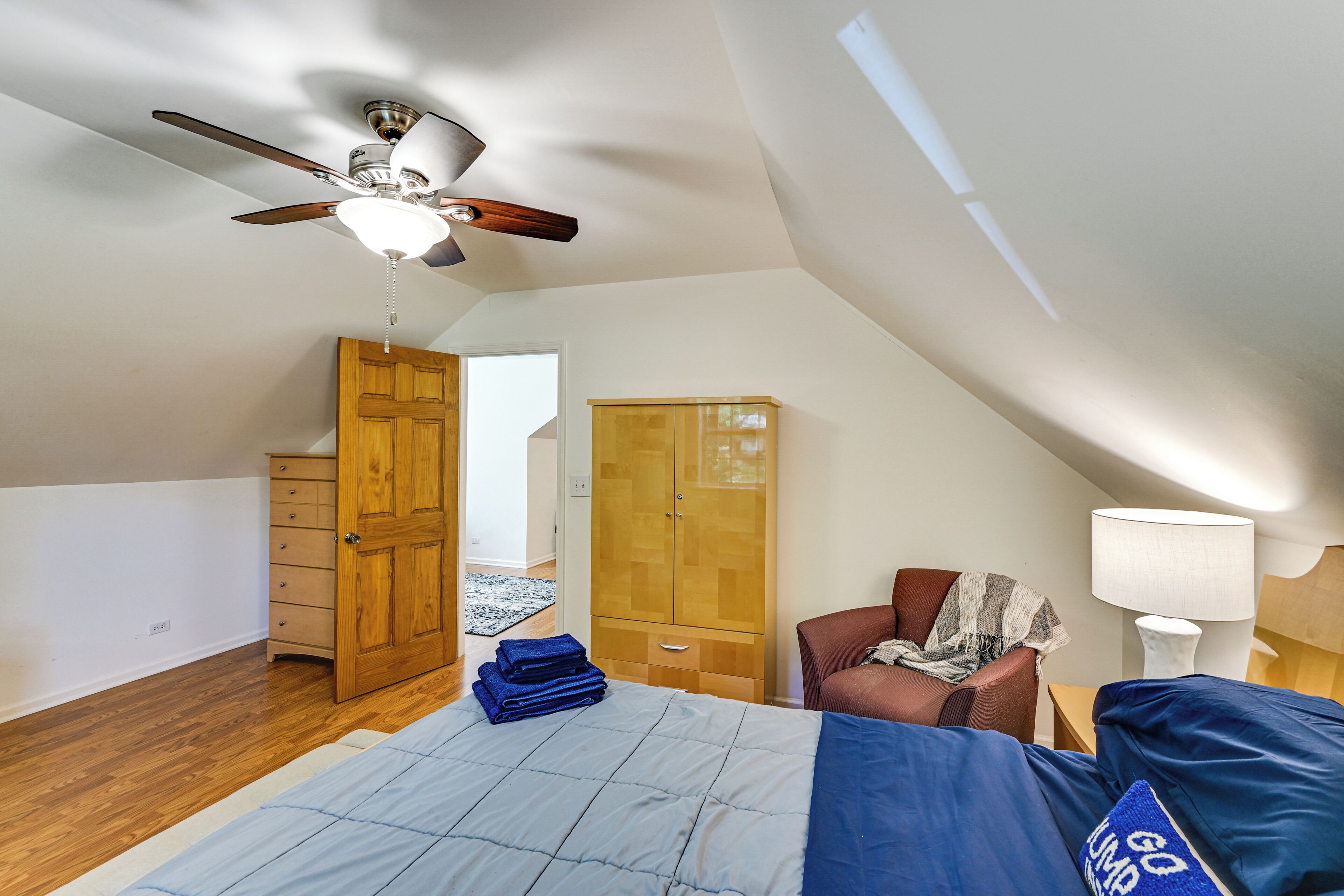 A bedroom with a bed, a ceiling fan, wooden wardrobe, and a chair.