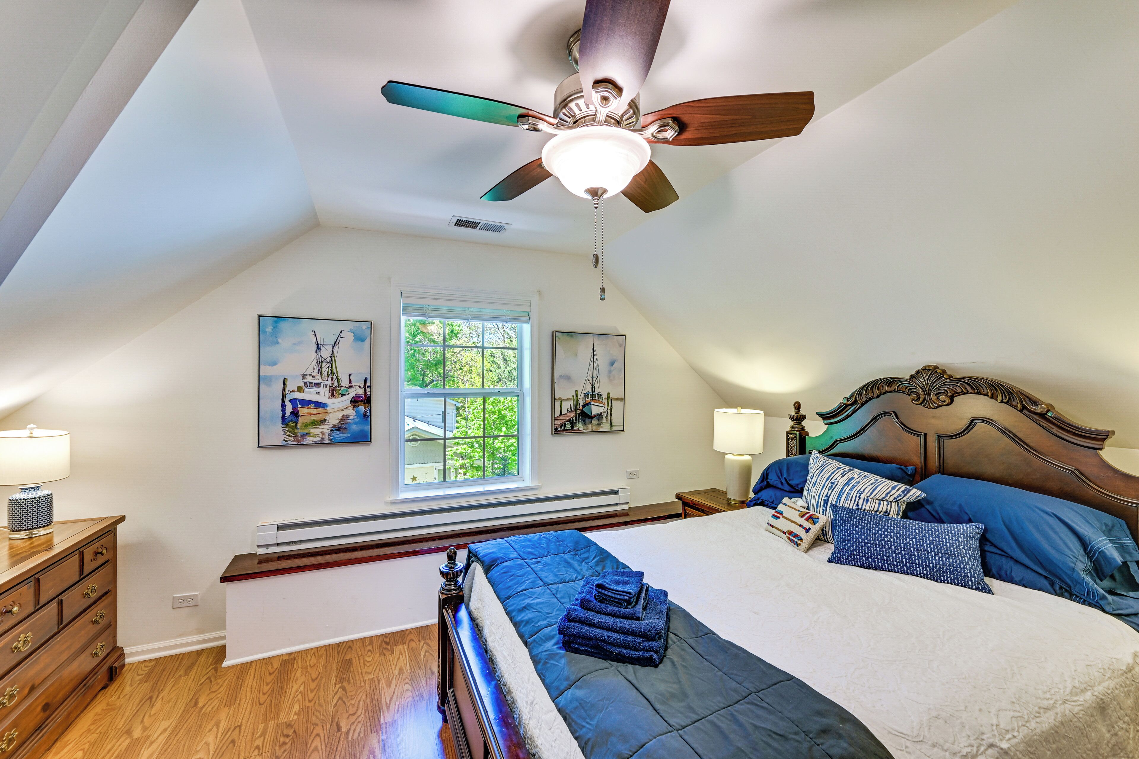 Ein Schlafzimmer mit schrägen Wänden, Deckenventilator, Bett mit blauer Bettwäsche, Nachttisch, Kommode und zwei an der Wand hängende Bilder.