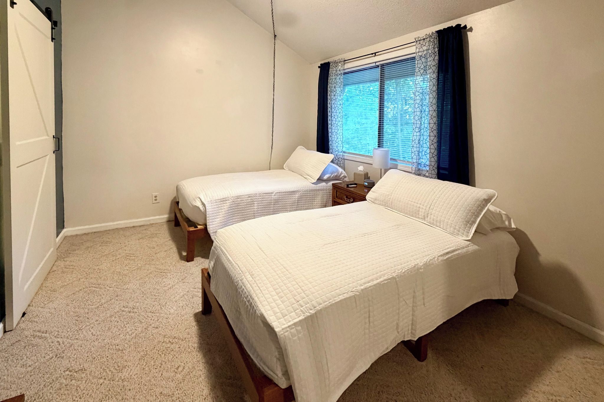 A bedroom with two beds, a window with blinds, a nightstand, and a lamp.