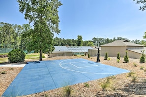 Ein Basketballplatz im Freien mit blauer Spielfläche, umgeben von Bäumen und einem Gebäude mit Satteldach.