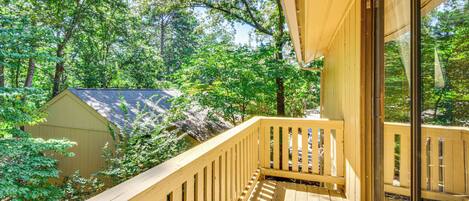 A wooden deck with a railing, overlooking a forested area with trees and a building in the background.