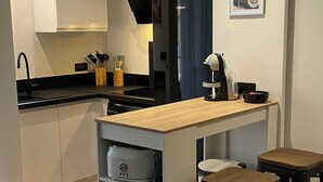 A modern kitchen with white cabinets, a black range hood, a Bosch refrigerator, and a coffee machine. There are two wooden stools and a framed picture on the wall. - Agréable Appartement de Type F3 (Argelès-sur-Mer)
