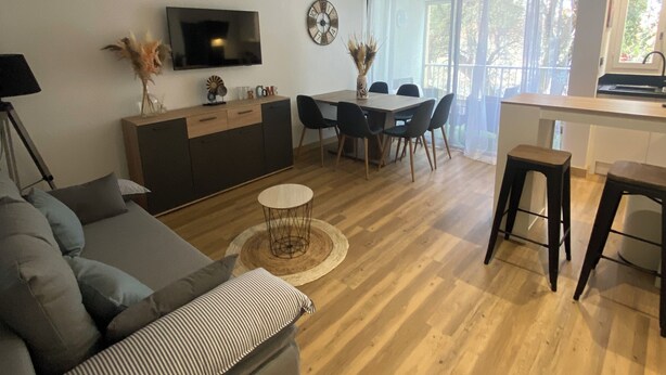 A modern living room with a sofa, a dining table with chairs, a TV mounted on the wall, and a kitchen area with a window. - Agréable Appartement de Type F3 (Argelès-sur-Mer)