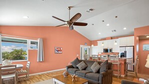 Un soggiorno con divano, tavolino e ventilatore a soffitto. È presente un angolo cottura con pensili e un tavolo da pranzo.