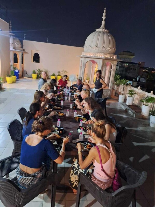 En gruppe mennesker spiser udendørs ved et rundt bord, med en pyntet pergola i baggrunden.