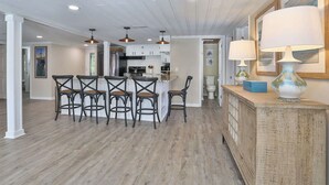 A spacious kitchen with a central island, bar stools, pendant lighting, and a wooden cabinet with a lamp. - The Clubhouse Murrells Inlet (Murrells Inlet)