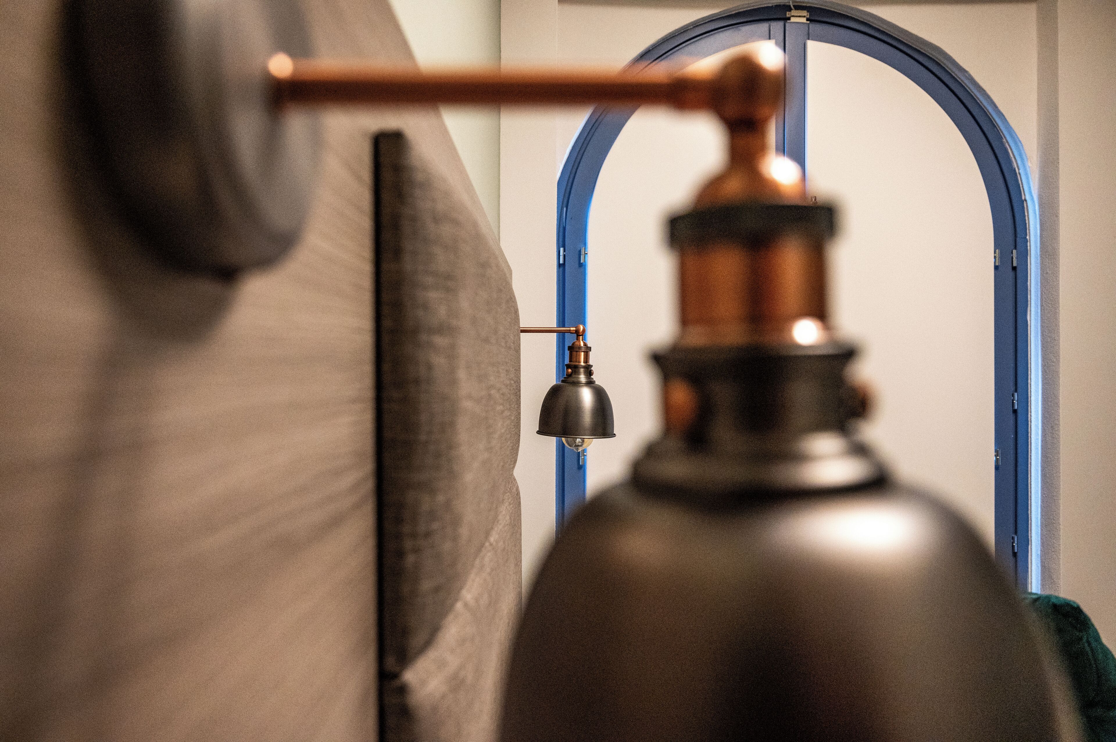 Een detail van een hoek van een kamer met een groot, donker, cilindrisch object, mogelijk een speaker of luchtreiniger, en een blauwe, gebogen deuropening.