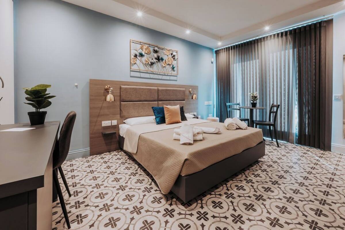 A modern hotel room with a large bed, a desk, and a chair. There is a plant on the desk and a painting on the wall.