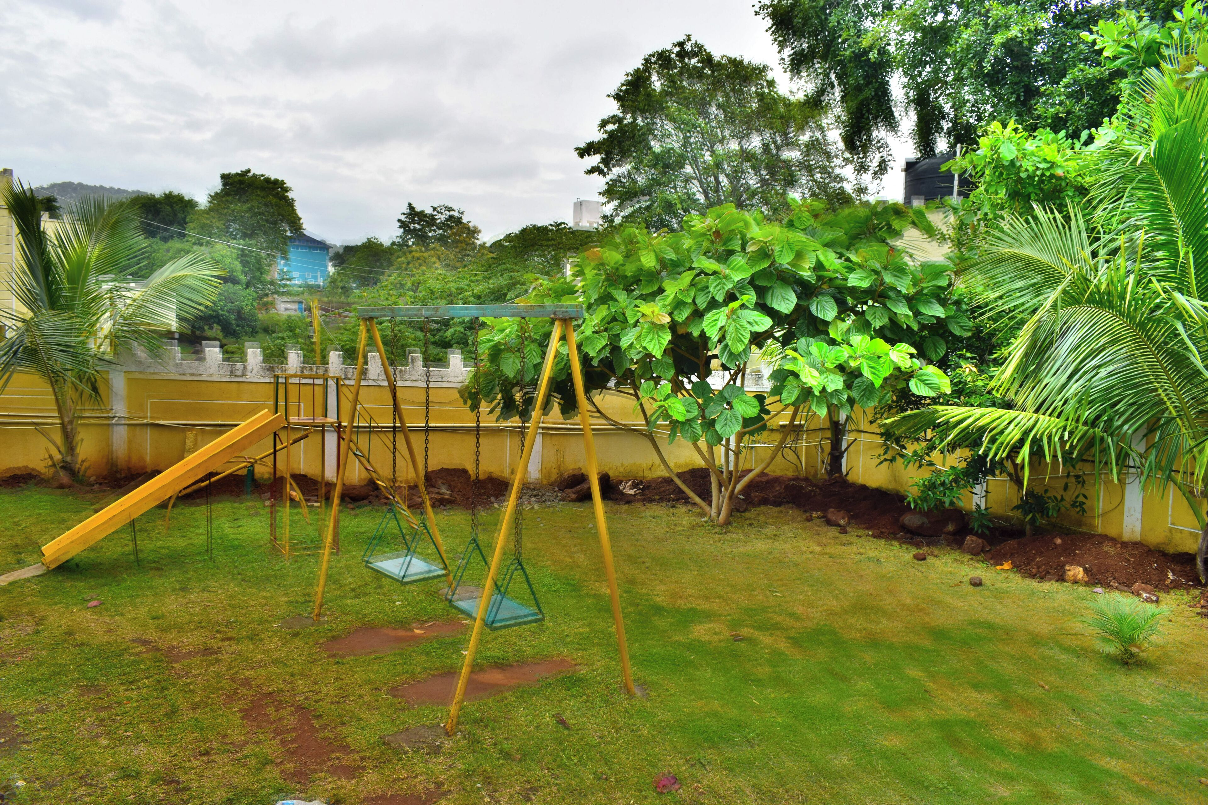 A playground with swings and a slide, surrounded by greenery and a yellow wall.