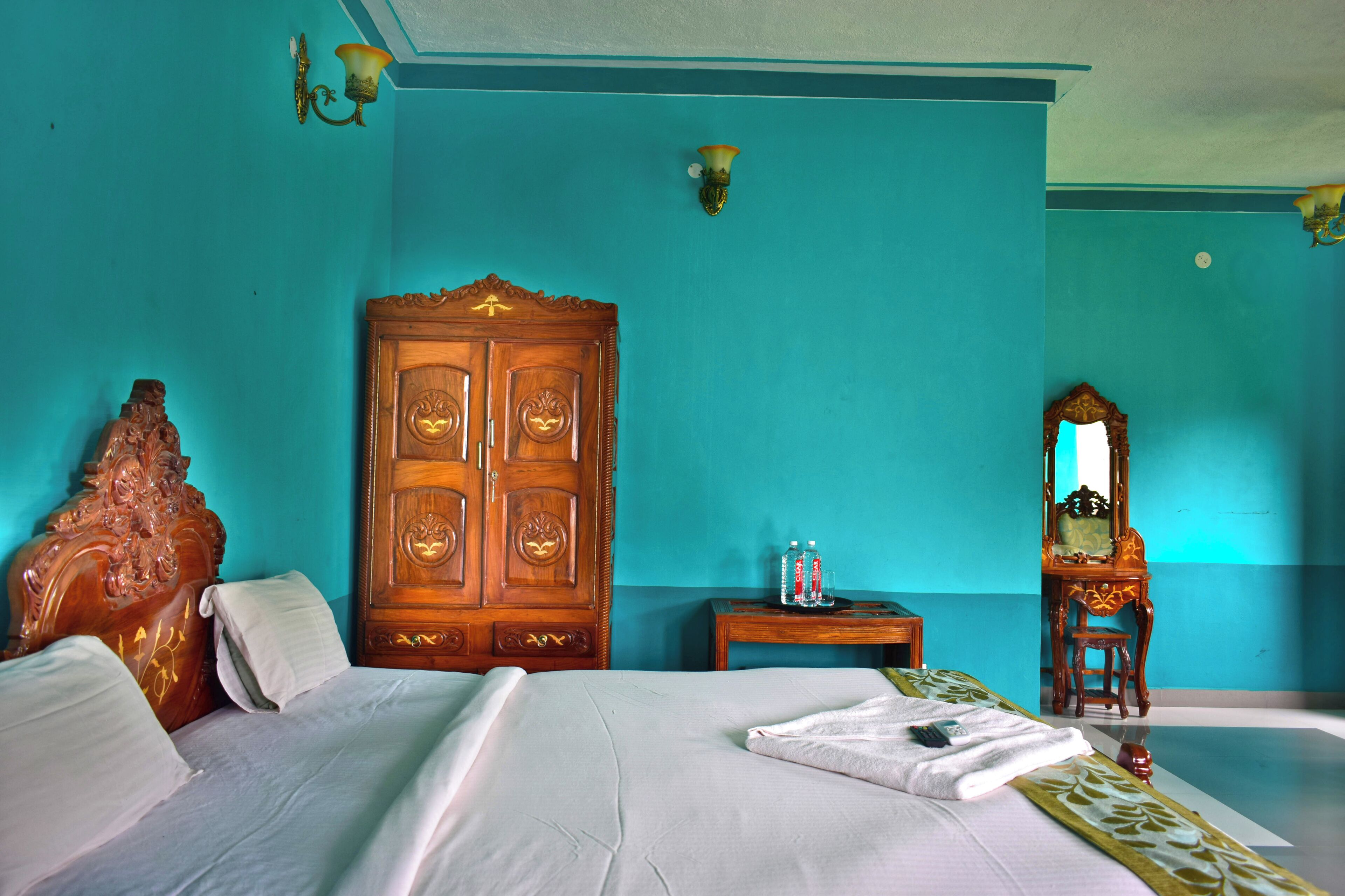 A room with a large wooden wardrobe, a bed with white linens, a small wooden table with a mirror, and teal walls.
