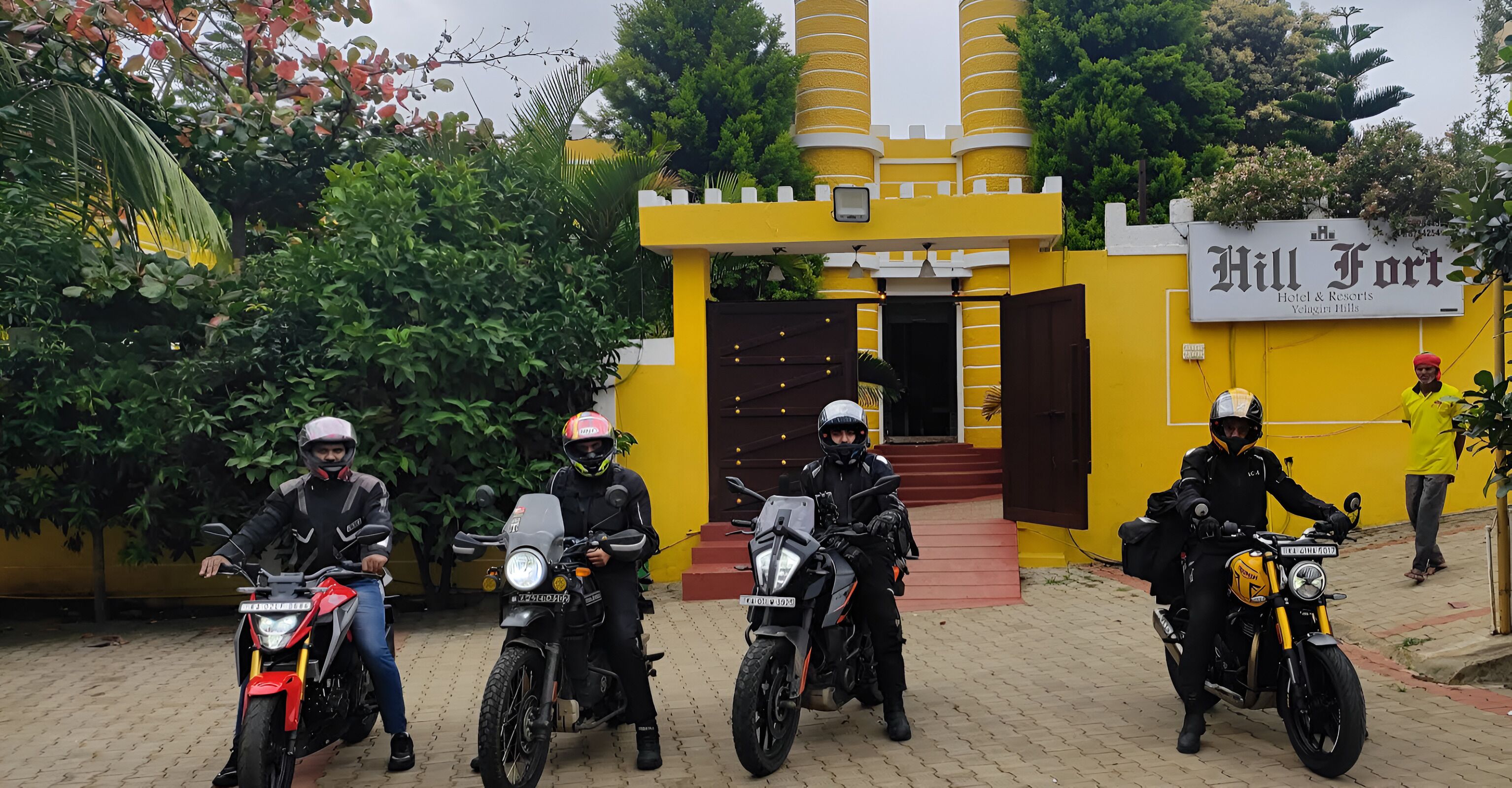 Cuatro motociclistas frente a un edificio amarillo con un letrero que dice 'Hill Fort Hotel & Resorts'.