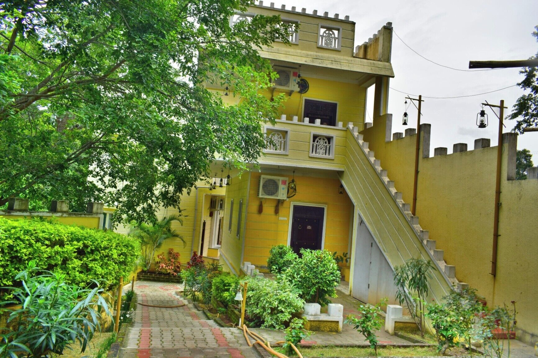 A two-story building with a yellow facade, a staircase leading to the upper floor, and a small garden area with various plants.