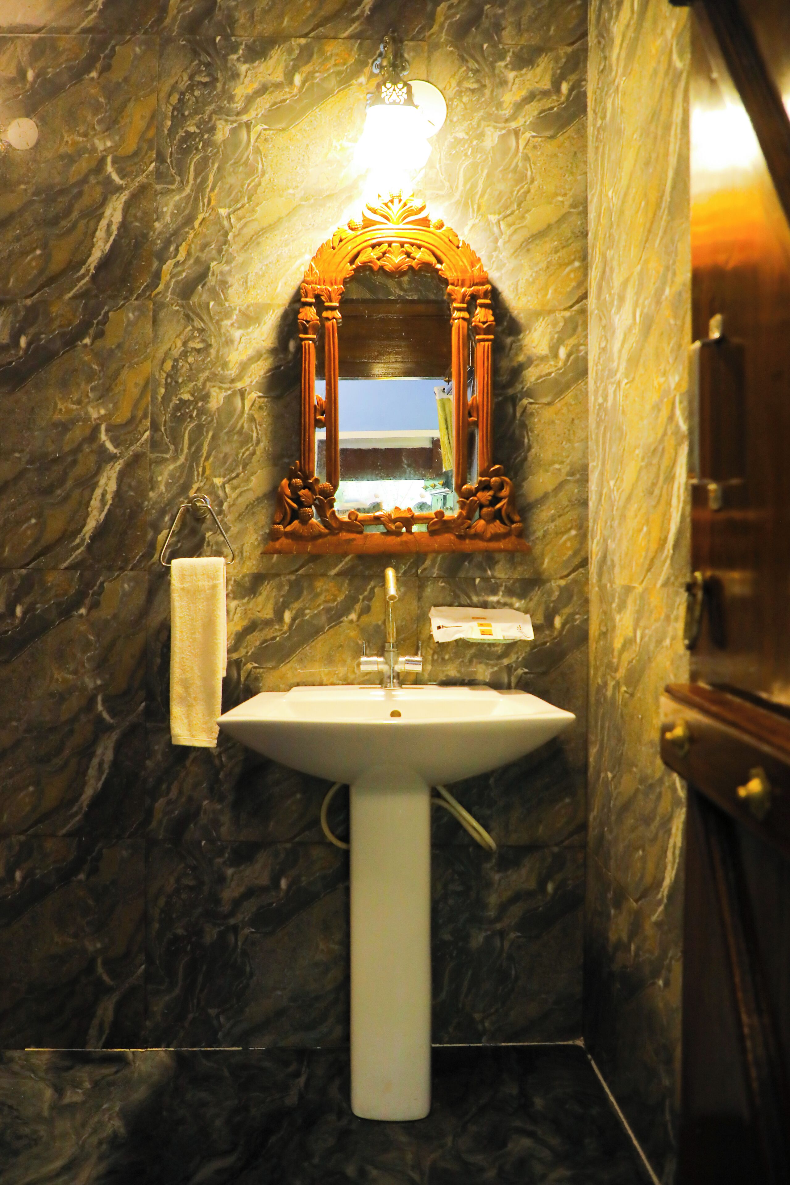 A bathroom with a marble wall, a sink, a mirror with an ornate frame, and a towel hanging on the wall.
