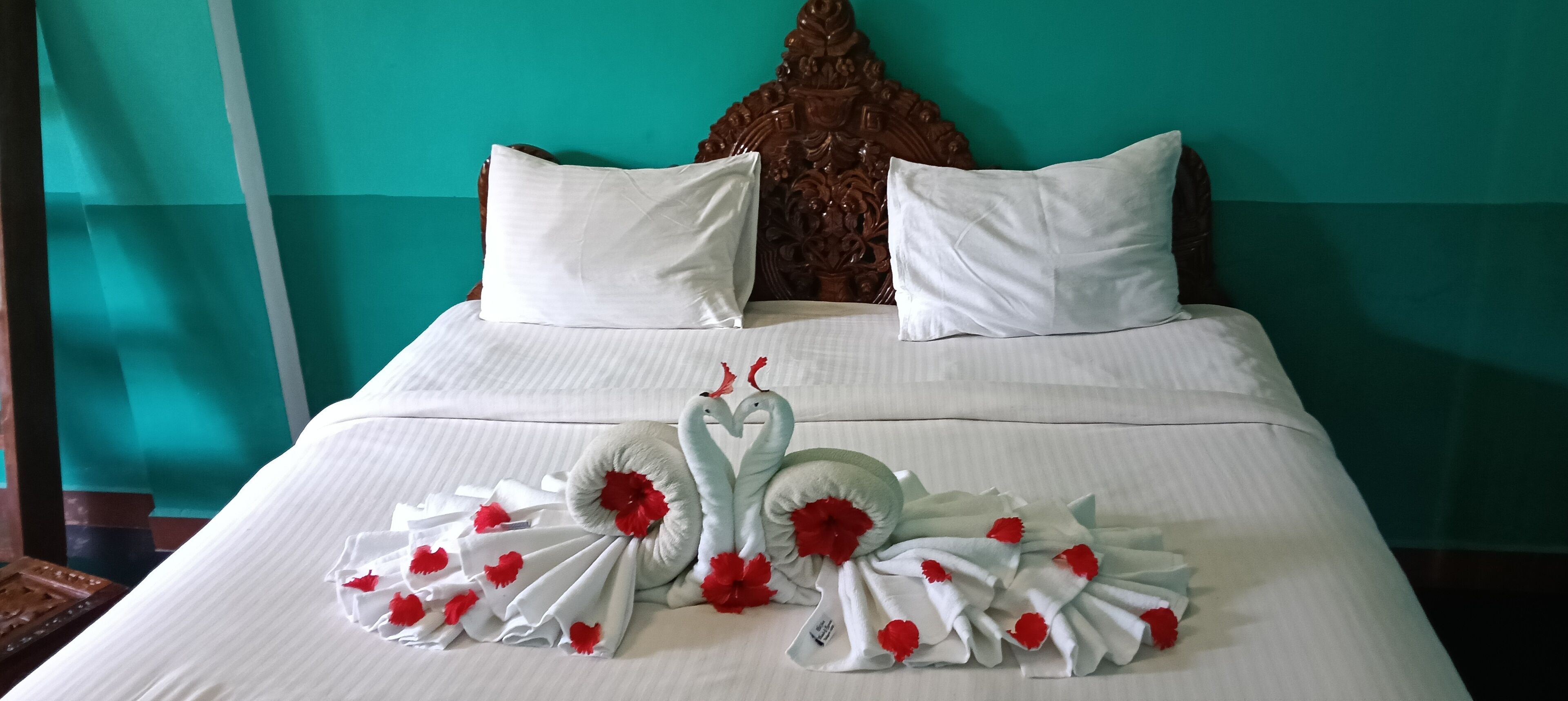 Een net opgemaakt bed met witte lakens en decoratieve handdoeken in de vorm van zwanen, versierd met rode bloemen, tegen een blauwgroene muur en houten hoofdbord.