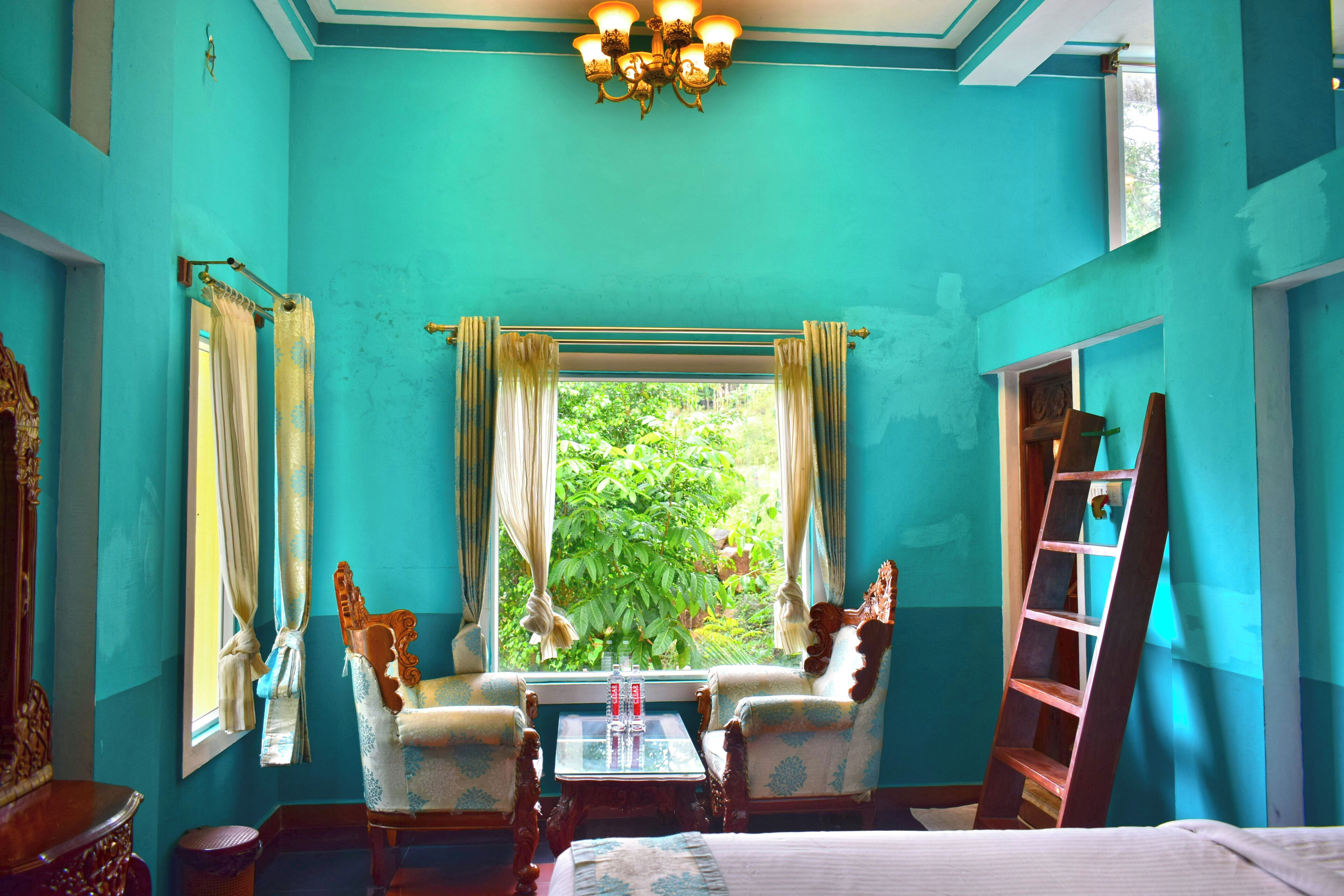 A room with teal walls, a chandelier, two ornate chairs, a small table, and a window with curtains.