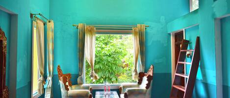 A room with teal walls, a chandelier, two ornate chairs, a small table, and a window with curtains.