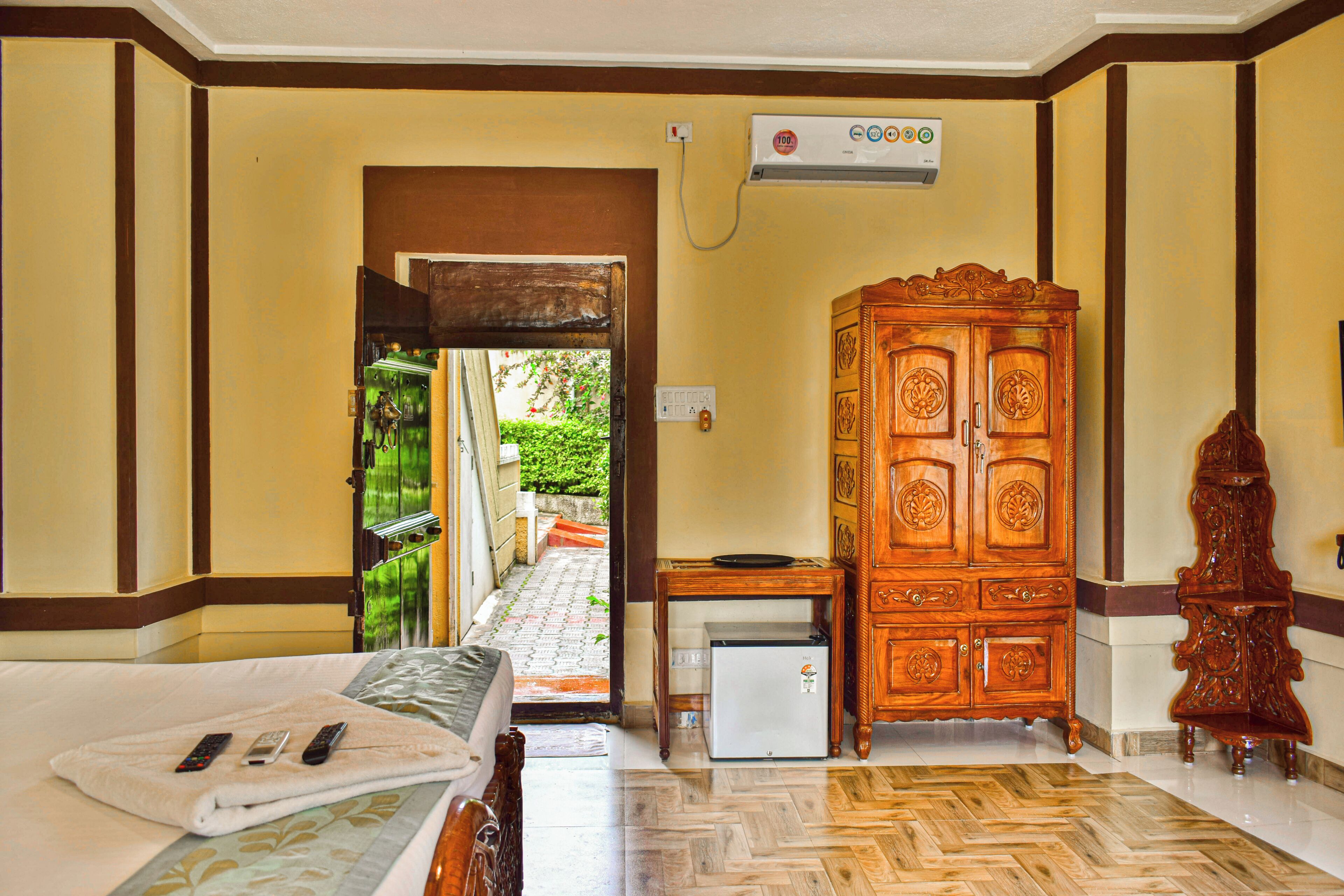 A hotel room with a bed, a small refrigerator, a wooden cabinet, and a door leading to a garden.