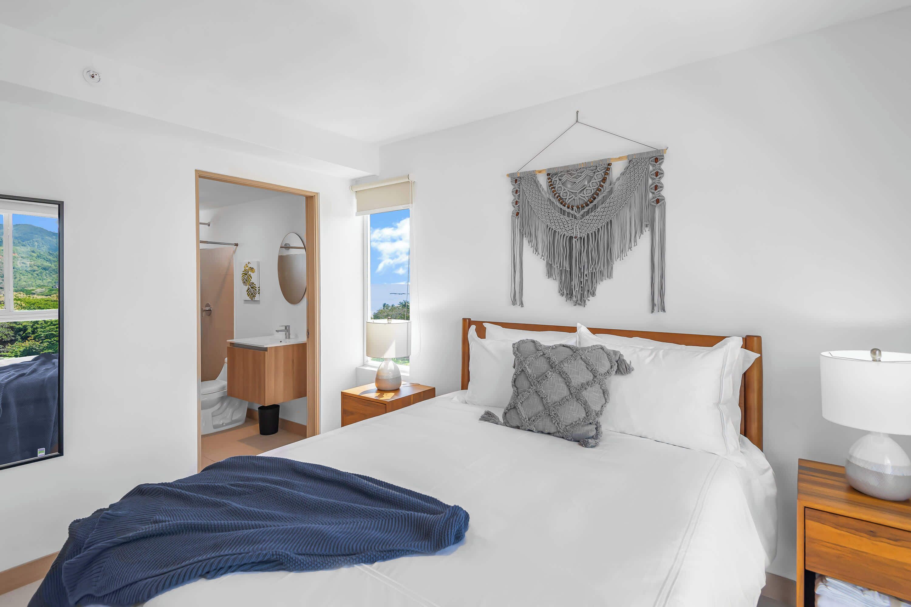 Un dormitorio con cama, mesita de noche con lámpara, decoración de macramé en la pared y vistas al exterior a través de la ventana.