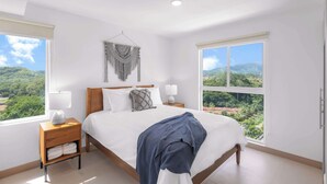 Une chambre avec un grand lit, deux tables de chevet, une décoration murale en macramé et une vue sur la verdure grâce à de grandes fenêtres.