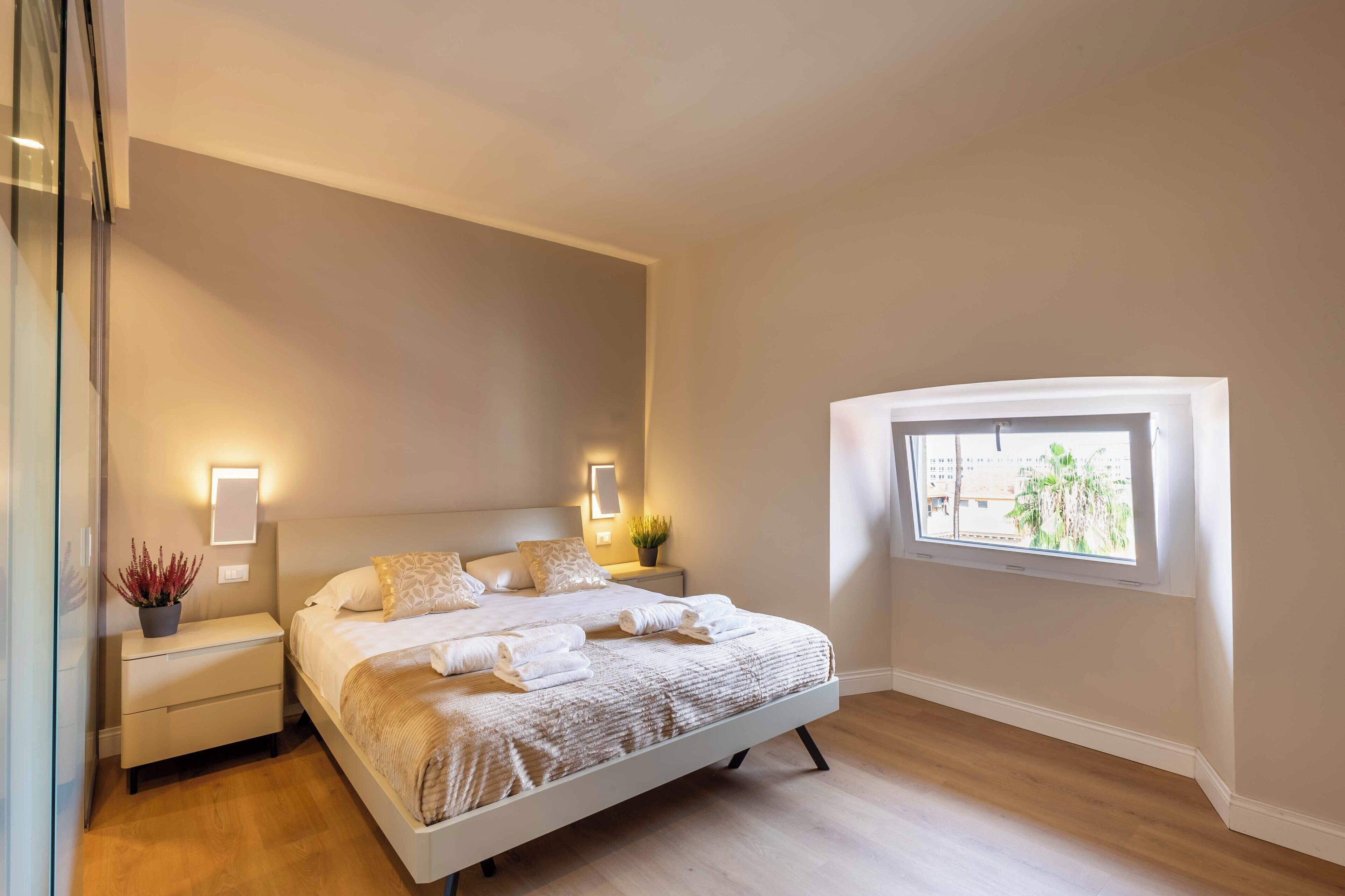 Une chambre moderne avec un grand lit, des tables de chevet, une commode, une fenêtre avec vue et des lampes fixées au mur.