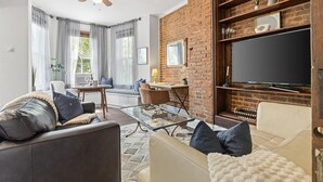 Una sala de estar con pared de ladrillo, un televisor de pantalla plana, una mesa de centro de vidrio y una zona de estar con sofá y sillones.