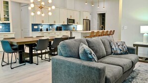 A modern kitchen with white cabinets, a central island, and a dining area with a grey sofa and chairs. - Angels Retreat @ Beach Club! (Gulf Shores)