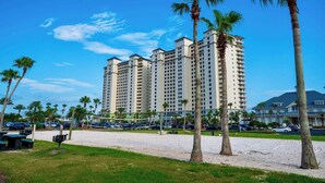 A beachside hotel with multiple high-rise buildings, palm trees, and a volleyball net. - Angels Retreat @ Beach Club! (Gulf Shores)