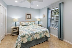 Un dormitorio con una cama, dos mesitas de noche con lámparas, un ventilador de techo, una ventana con persianas y un cuadro en la pared.