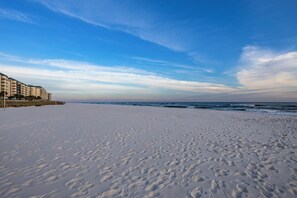 Une plage parsemée d'empreintes, un ciel dégagé et des bâtiments en arrière-plan.