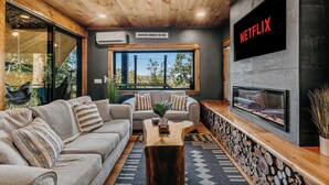 Un soggiorno moderno con camino, un grande schermo TV che mostra 'NETFLIX' e un'area salotto accogliente con vista sull'esterno.