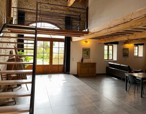 A spacious room with a high ceiling, wooden beams, a mezzanine level, and large windows allowing natural light. - 3-star holiday cottage in a barn with swimming poo (Fiac)