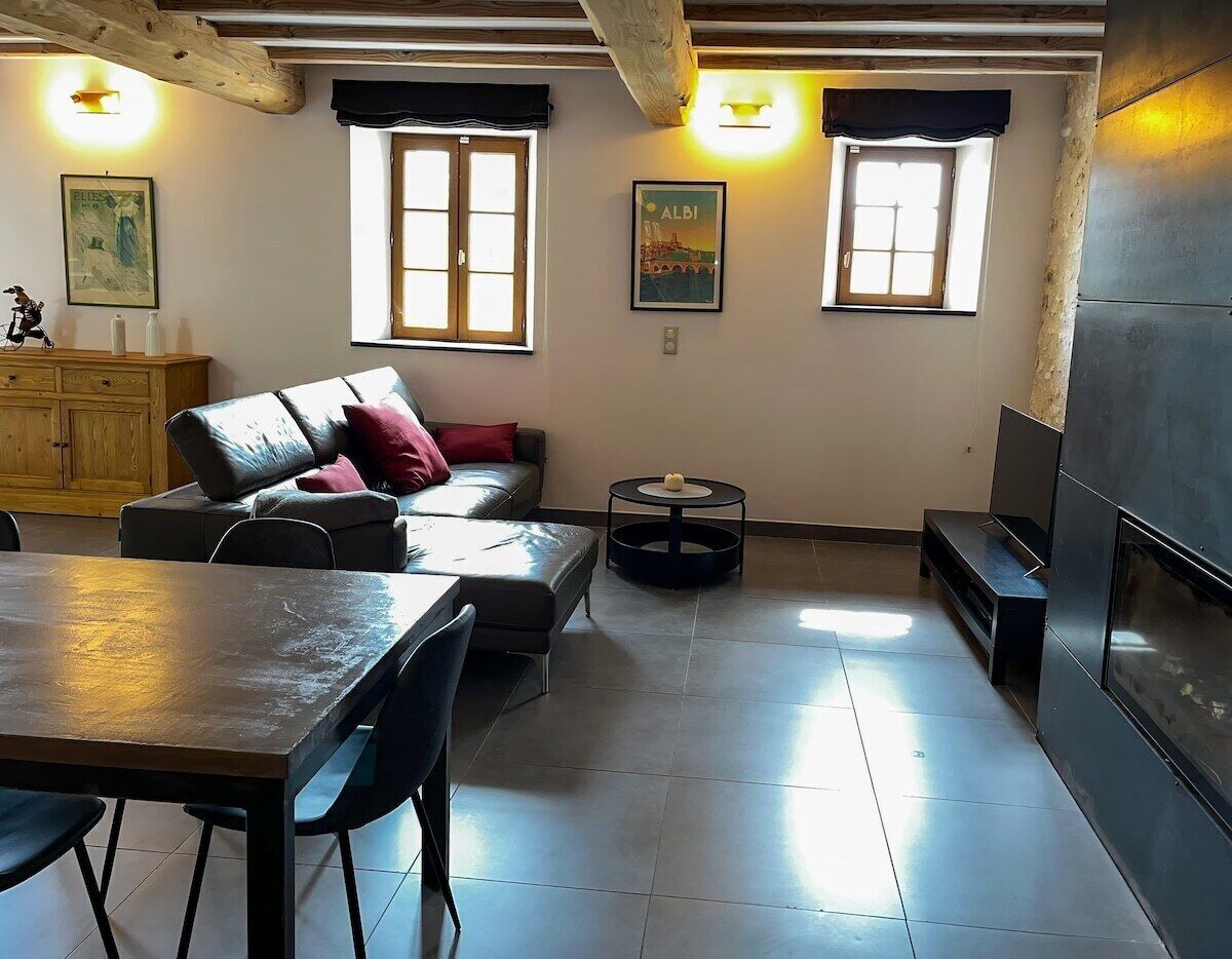 A modern living room with a sofa, a dining table, and a fireplace. There are two windows with curtains, a wooden cabinet, and a small round table.
