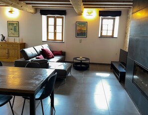 A modern living room with a sofa, a dining table, and a fireplace. There are two windows with curtains, a wooden cabinet, and a small round table.
