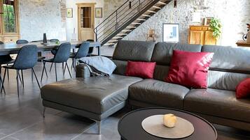 A modern living room with a sectional sofa, a dining area with chairs, and a staircase against a stone wall.