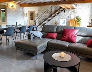 A modern living room with a sectional sofa, a dining area with chairs, and a staircase against a stone wall.