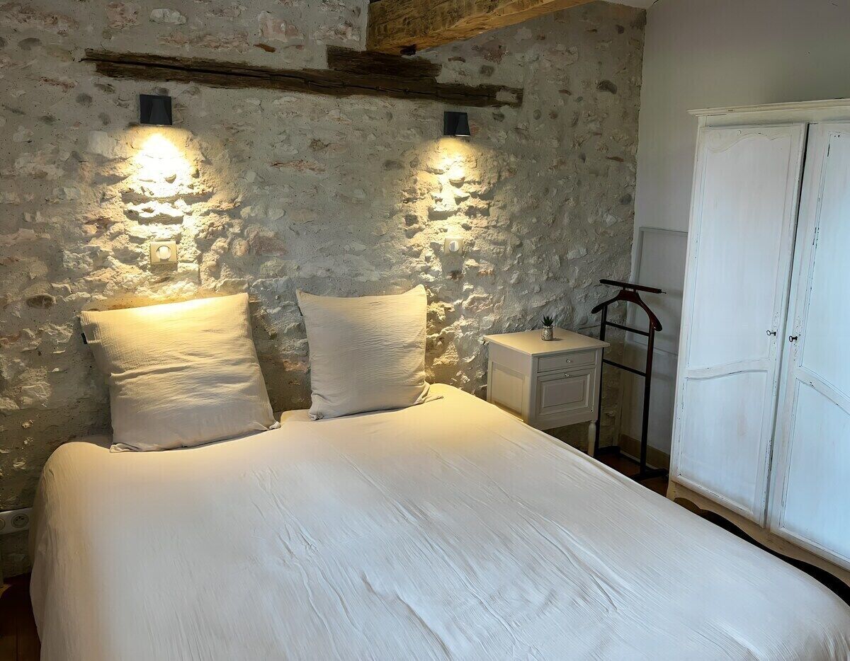 Un llit amb dos almofadons, una tauleta de nit i un armari a una habitació amb parets de pedra i bigues de fusta.