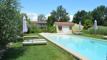A swimming pool with lounge chairs and umbrellas, surrounded by greenery and a building with a tiled roof.