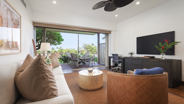 A modern living room with a sofa, coffee table, and wicker chairs. A TV is mounted on the wall, and there is a sliding glass door leading to an outdoor area with a table and chairs. - Luxury 3BR Ka 'Ulu Villa 129B at Hualalai – Island Elegance & Golf Views (Kailua Kona)
