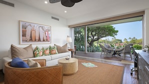 A spacious living room with a large window, a sofa with floral cushions, a coffee table, and a dining area with wicker chairs. - Luxury 3BR Ka 'Ulu Villa 129B at Hualalai – Island Elegance & Golf Views (Kailua Kona)