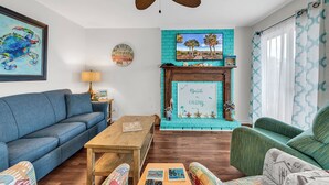 A living room with a blue sofa, a wooden coffee table, a green armchair, a fireplace with a beach-themed sign, and a ceiling fan. - Gorgeous modern condo with unmatched view looking out over the pool! (Surfside Beach)