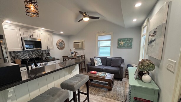 A modern kitchen with a central island, a dining area with a sofa and coffee table, and a living room with a couch and a bookshelf. - The Nautical Nest-Belle of Maine (Wells)