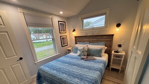 A bedroom with a large bed, two bedside tables, a window with blinds, and a view of a house outside. - The Nautical Nest-Belle of Maine (Wells)