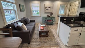 A modern living room with a dark brown sofa, a wooden coffee table, and a kitchen area with white cabinets and a black countertop. - The Nautical Nest-Belle of Maine (Wells)