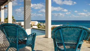 A view of a beachside pool area with two wicker chairs facing the ocean, under a white pergola.