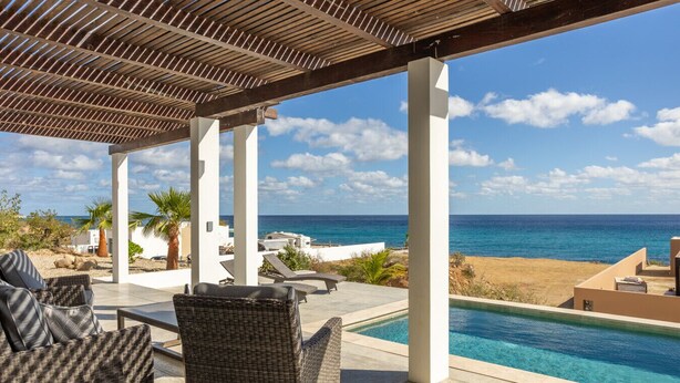 A covered outdoor area with a pool and lounge chairs, overlooking a beach and the ocean.